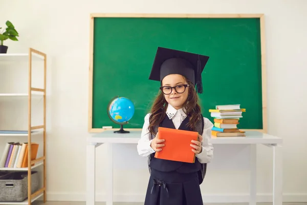 Πίσω στο σχολείο. Μια χαριτωμένη μαθήτρια με καπέλο αποφοίτησης με βιβλία στέκεται στο φόντο μιας τάξης σε ένα σχολείο.. — Φωτογραφία Αρχείου