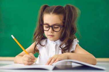 Okul konseptine geri dönelim. Gözlüklü zeki bir kız sınıfta bir masada otururken kalemle bir not defteriyle yazar..