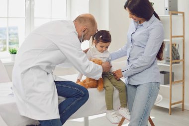 Steteskoplu kıdemli doktor, annesinin hastanedeki hastalarına bakan küçük bir kızı dinliyor..
