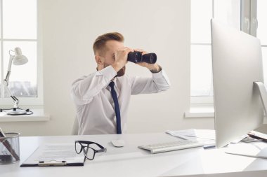 A businessman looks through binoculars while sitting at a table with a computer in the office.