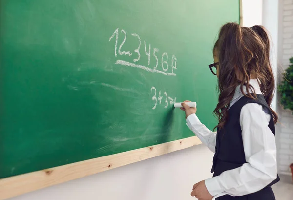 Terug naar school. Een schoolmeisje met een bril schrijft op een schoolbord in een klaslokaal op een lezing. — Stockfoto