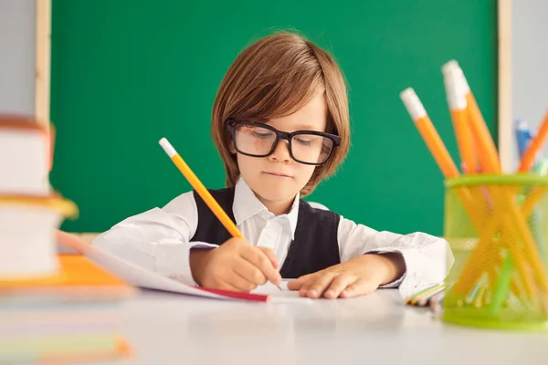 A estudar. Um estudante inteligente com óculos escreve um trabalho de professor em um caderno em uma mesa na sala de aula . — Fotografia de Stock