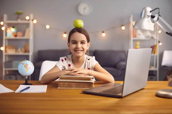 De volta à escola. Estudante feliz com maçã ina cabeça sorri enquanto sentado à mesa com laptop em casa . — Fotografia de Stock