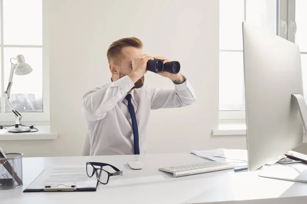 A businessman looks through binoculars while sitting at a table with a computer in the office. — ストック写真