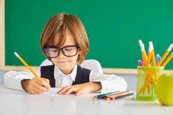 Повернутися до школи. Розумний хлопчик, школяр пише олівцем, сидячи за столом на фоні шкільної дошки . — стокове фото
