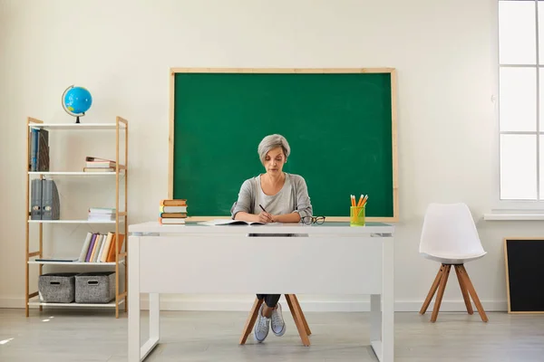 Professeur mature sérieux assis au bureau et écrivant en classe. Professeure se préparant pour la conférence à l'école — Photo