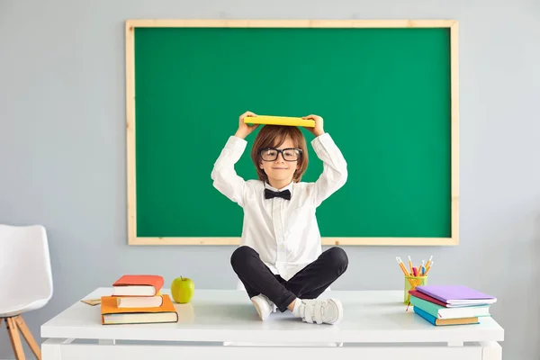 La educación. De vuelta a la escuela. Divertido colegial sostiene un libro sobre su cabeza mientras está sentado en un escritorio en la escuela — Foto de Stock