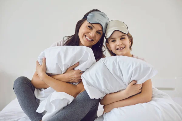 Hari ibu bahagia. Ibu dan anak dalam topeng untuk tidur tersenyum sambil duduk dengan bantal melihat kamera di tempat tidur di kamar. — Stok Foto