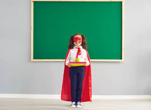 Écolière en costume de super-héros avec des livres debout sur le fond du tableau noir de l'école. — Photo