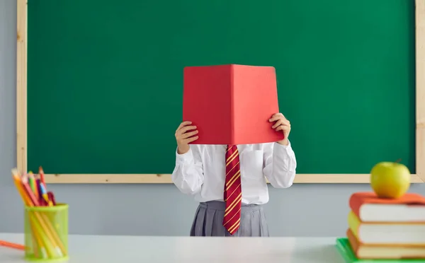 Tillbaka till skolan. En skolflicka läser en bok, täcker ansiktet med en svart tavla i klassrummet.. — Stockfoto