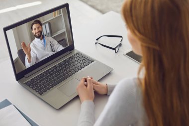 Çevrimiçi doktor danışmanlığı. Erkek doktor dizüstü bilgisayardan el sallıyor. Evde oturan bir kız hasta için video danışmanlığı yapıyor..