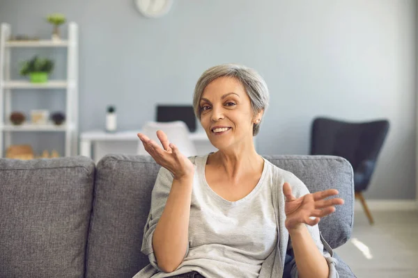 Heureuse femme âgée avec les cheveux gris dit en regardant la caméra tout en étant assis sur un canapé à la maison. — Photo