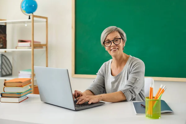 Escuela de profesores universidad en línea. Mujer profesora sénior habla con los estudiantes tiene chat web utilizando ordenador portátil sentado en el escritorio en el aula . —  Fotos de Stock
