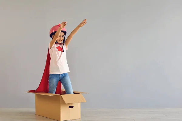 Un enfant en costume de super-héros leva la main debout dans une boîte en carton un fond gris. — Photo
