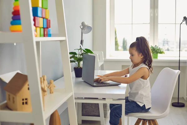 Educación en línea de los niños. Niña seria está escribiendo texto portátil mientras está sentado en una mesa en casa . —  Fotos de Stock