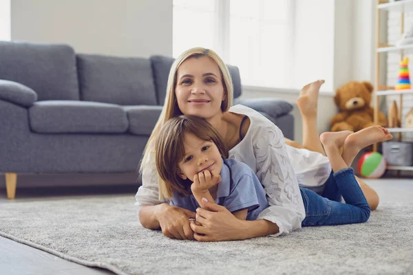 Liebevolle Mutter umarmt ihren Sohn, während er auf dem Teppich im Wohnzimmer liegt. Eltern und Kind umarmen sich zu Hause. Familienbande — Stockfoto