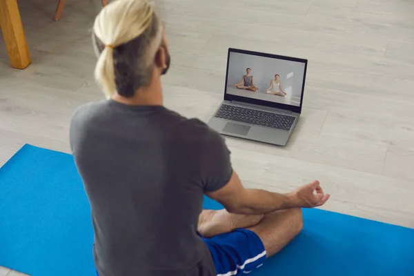 Yoga en línea en casa. Hombre joven siguiendo clases de yoga en línea y haciendo su meditación matutina o ejercicios de respiración en la esterilla en casa — Foto de Stock