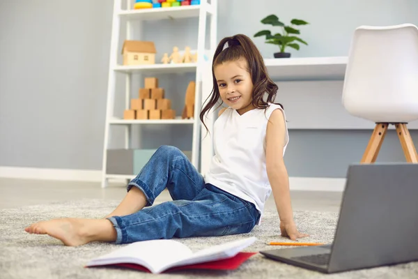 Educación en línea. Retrato de colegiala sentada en el suelo cerca de la computadora portátil y portátil. Niño inteligente estudiando en casa —  Fotos de Stock