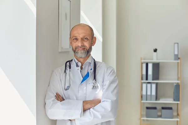 Oude dokter met gekruiste armen in een modern kliniek kantoor. — Stockfoto