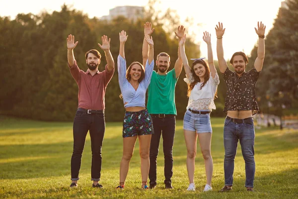 Een groep vrienden hief vrolijk hun hand op bij een picknickfeest in een zomerpark bij zonsondergang.. — Stockfoto