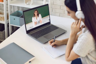 Çevrimiçi öğretmen. Kulaklıklı kız öğrenci, üniversite öğretmeninin videolu sohbet konferansını dinliyor. Evde bilgisayarı var..
