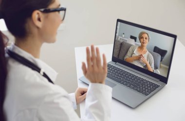 Doktor kız, yaşlı bir kadına danışmak için elini sallıyor. Klinikteki bir masada dizüstü bilgisayar kullanıyor..