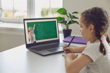 Online çocuk eğitimi. Küçük kızın videolu konferans dizüstü bilgisayarı ve evde oturan bir öğretmeni var..
