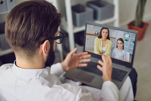 Male doctor therapist gives recommendations for treating a sick child using a laptop video webcam while sitting in a clinic office. — Stock Photo, Image