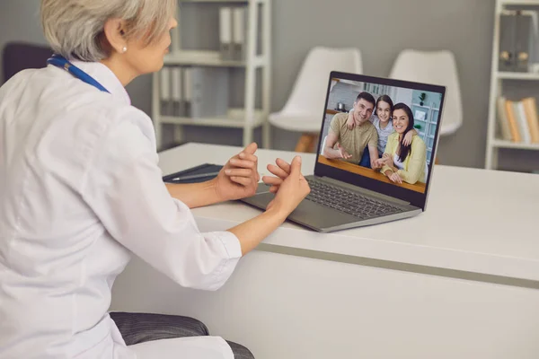 Лікар онлайн. Лікар з сірим волоссям консультує сім'ю і проводить конференцію з відео чату ноутбука в клінічному офісі . — стокове фото