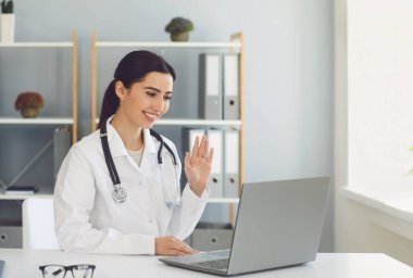 Bir hastaya el sallayan İspanyol kadın doktorun klinikteki bir masada video konferans dizüstü bilgisayarı var..