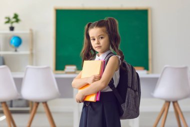 Okul konseptine geri dönelim. Elinde kitaplarla küçük bir kız öğrenci, ilkokulda sınıfın arka planında duruyor..