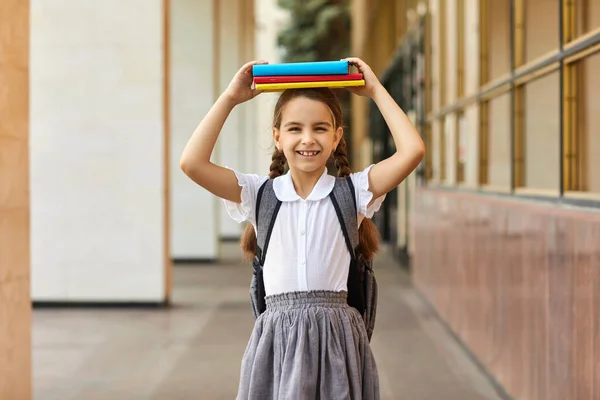 Je retourne à l'école. Écolière en uniforme tenant un tas de livres sur sa tête près du bâtiment de l'école. — Photo