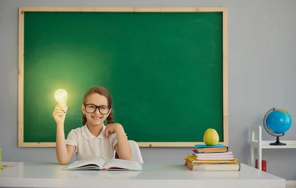 Una colegiala sostiene en su mano una lámpara con luz sentada en un escritorio de la escuela . — Foto de Stock