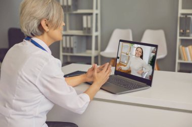 Hastanedeki genç bir hastayla görüntülü konuşma yapan kıdemli doktor. Doktor ofisteki dizüstü bilgisayarda müşteriyle konuşuyor.