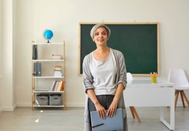 Sınıftaki öğretmenim. Gri saçlı bir kadın elinde not defteri olan bir öğretmen sınıfın arka planında duruyor..