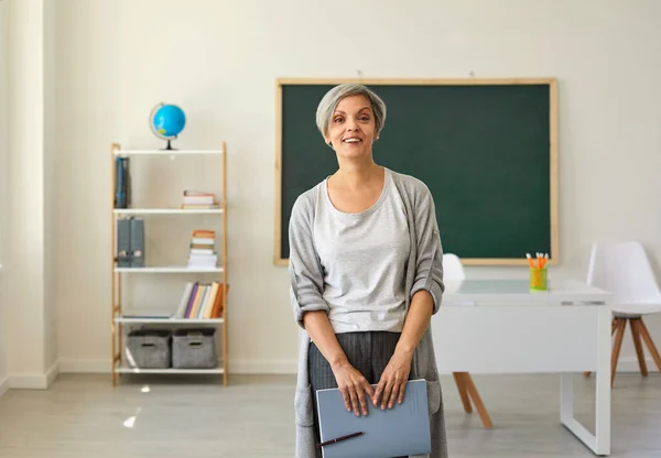 Le professeur dans la salle de classe. Une femme aux cheveux gris un professeur avec un cahier à la main debout sur le fond de la classe. — Photo