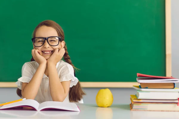 Concept back to school. Funny schoolgirl with glasses smiling while sitting at the table with books in the classroom.