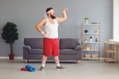 Spor kıyafetli komik şişman adam evde duran kaslarını gösteriyor.