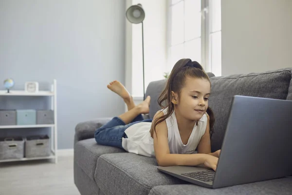 Holčička ležící na gauči s laptopem a dívající se doma na online lekci. Dítě se vzdělávacím kurzem na webu — Stock fotografie