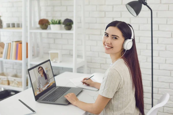 Lächelndes junges Mädchen mit Kopfhörern, das online mit seinem Chef oder Lehrer kommuniziert und sich Notizen im Copybook macht, drinnen — Stockfoto