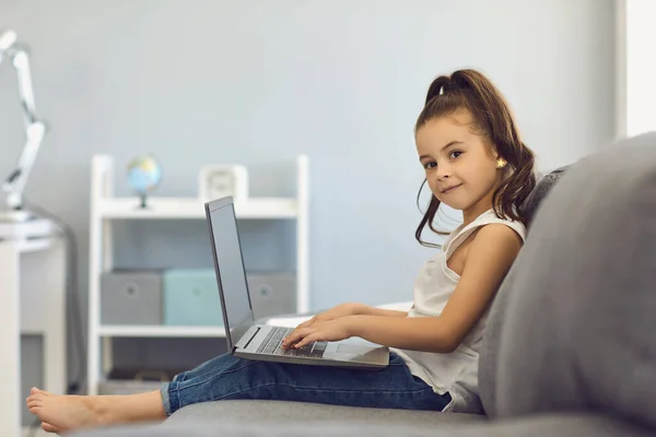 Chica utiliza un ordenador portátil mientras está sentado en el sofá en la habitación en casa . — Foto de Stock