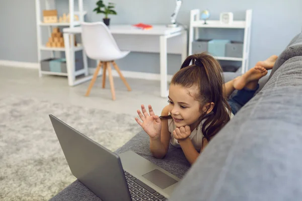 Маленькая девочка, лежащая на диване с ноутбуком и смотрящая видео дома. Обучение детей онлайн. — стоковое фото
