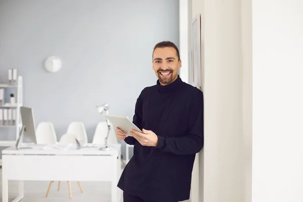 Un tipo positivo sonriendo de pie con una tableta en la mano en la oficina en el lugar de trabajo . — Foto de Stock