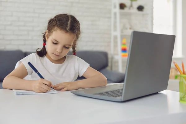 Online oktatás és otthoni oktatás. Okos kislány jegyzetel a laptop előtt a webes lecke alatt — Stock Fotó