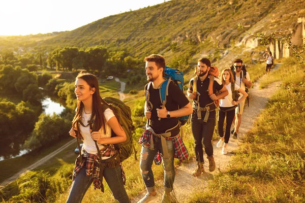 Jóvenes con mochilas caminando por senderos estrechos en las montañas. Grupo de turistas alegres haciendo senderismo fuera en verano —  Fotos de Stock