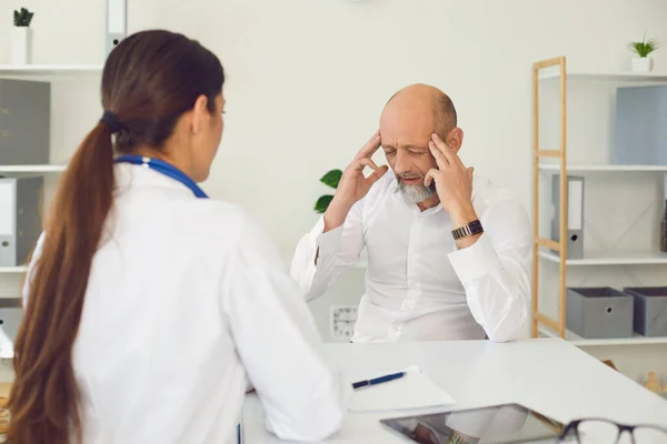 Hoofdpijn. Patiënt senor vertelt de arts hoofdpijn symptomen zittend op een stoel in een kliniek kantoor. — Stockfoto