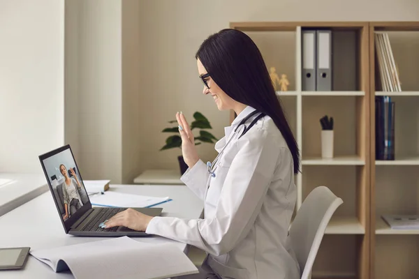 사무실의 한 여의사가 랩탑으로 환자에게 손을 흔들고 있습니다. 온라인 의사는 환자의 나이든 여성에게 화상 통화를 사용하여 원격 상담을 한다. — 스톡 사진