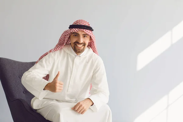Άραβας άνδρας χαμογελά ενώ κάθεται σε μια καρέκλα σε ένα γκρι φόντο. — Φωτογραφία Αρχείου