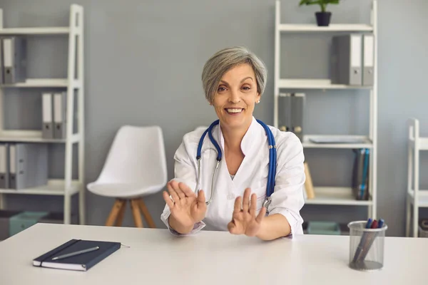 Femme âgée souriante médecin parle dans une webcam ordinateur en ligne alors qu'il est assis dans un bureau de clinique. — Photo