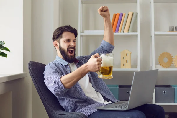 Evde elinde bira kupasıyla dizüstü bilgisayarda futbol izleyen genç adam. Spor taraftarı içeride futbol takımı için tezahürat yapıyor. — Stok fotoğraf